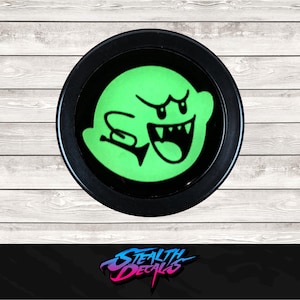 May include: A round, black decal featuring a green cartoon ghost with a wide open mouth and sharp teeth. The ghost has a black outline and is set against a black background. The decal is on a light wood grain surface.