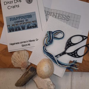 May include: A cross stitch kit with blue, white and yellow embroidery floss, a white Aida cloth and a pair of black scissors. The kit includes instructions for a cross stitch pattern that reads "Happiness comes in waves".