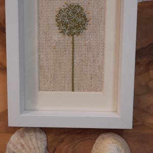 May include: A framed cross-stitch embroidery of a green dandelion with white seeds on a beige linen background.