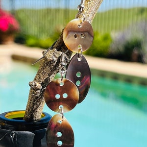 May include: A pair of earrings made from three brown shell buttons. The top button is round, the middle button is round, and the bottom button is oval shaped. The earrings are hanging from a silver wire.