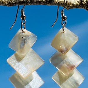 Puede incluir: Un par de pendientes de concha blanca con un diseño geométrico en capas. Los pendientes están hechos de tres piezas cuadradas de concha, apiladas una encima de la otra. Los pendientes están unidos a ganchos plateados.
