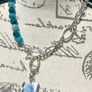 May include: A silver chain necklace with a large, rectangular pendant. The chain is made of thick, oval links. The pendant is smooth and has a slightly textured surface. The necklace is displayed on a white fabric background.