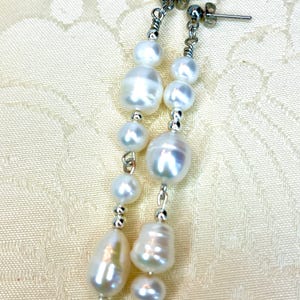 Könnte beinhalten: Ein Paar silberne Ohrhänger mit weißen Perlen-Tropfen. Die Ohrringe haben einen silbernen Hakenverschluss.