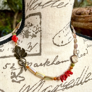 May include: A multi-colored necklace with a leafy bronze clasp. The necklace features red, orange, brown, and white beads, as well as a large, square, iridescent shell bead.