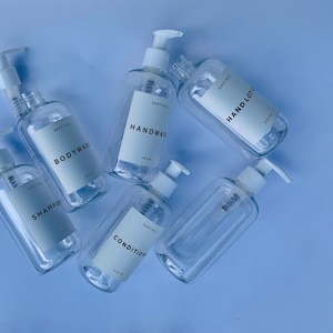 Puede incluir: Cinco botellas de plástico transparente con bombas, cada una etiquetada con un nombre de producto diferente: "Handwash", "Bodywash", "Shampoo", "Conditioner" y "Hand Lotion". Cada botella está etiquetada con "East Hill" y el volumen "500 ml".