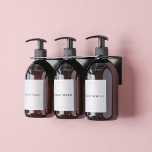 May include: Three black pump bottles with labels that read "Shampoo", "Conditioner", and "Body Wash" are mounted on a black wall-mounted dispenser. The bottles are filled with a dark liquid and have a white label with black text.