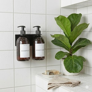 May include: Two amber glass pump bottles mounted on a black wall bracket, labelled Shampoo and Conditioner in black text on white rectangular labels. The bottles feature black pump dispensers and are positioned against a white tiled wall.