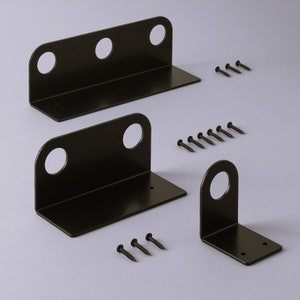 Pode incluir: Três suportes de prateleira de metal preto com furos para parafusos. Os suportes são de tamanhos diferentes e estão dispostos em forma de escada sobre uma superfície cinza. Existem vários parafusos pretos pequenos espalhados ao redor dos suportes.