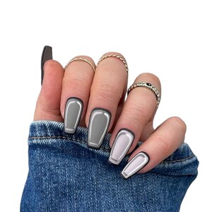Könnte beinhalten: Eine Hand mit Sarg-förmigen Kunstnägeln in Grau und Weiß mit schwarzem Umriss. Die Hand ist mit goldenen Ringen geschmückt und trägt eine Jeansjacke.