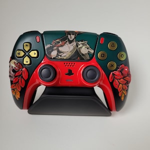May include: Custom-designed video game controller featuring a red and black color scheme with gold buttons. The controller has a green top with a graphic of a character. Decorative floral and skull elements are on the sides. The controller is on a black stand.
