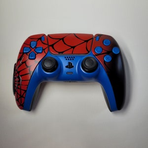 May include: A custom PlayStation 5 controller featuring a Spider-Man design. The controller has a red web pattern on the top, blue accents, and black grips. The buttons are blue, and the joysticks are black.