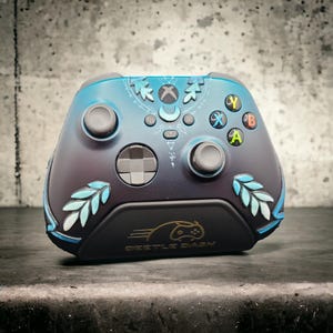 Könnte beinhalten: Benutzerdefinierter Xbox-Controller mit einem Farbverlauf in Blau und Schwarz. Der Controller verfügt über Blatt- und Mondakzente sowie farbige Tasten. Auf der Unterseite des Controllers steht "Beetle Dash" in Gold.