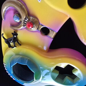 May include: Close-up of a colorful, abstract mask with a glossy finish. The mask features a black cat figurine, gold accents, and embedded gemstones. The color gradient transitions from yellow to pink and blue.