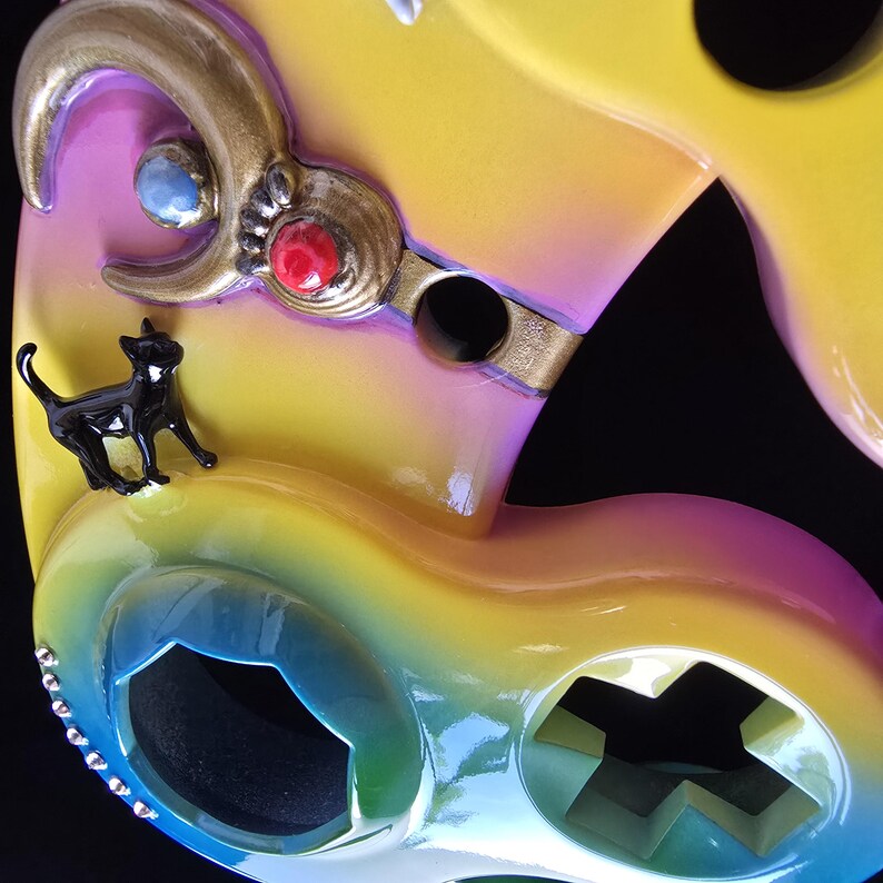 May include: Close-up of a colorful, abstract mask with a glossy finish. The mask features a black cat figurine, gold accents, and embedded gemstones. The color gradient transitions from yellow to pink and blue.