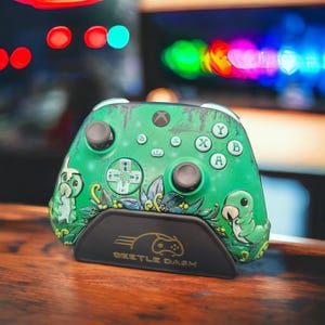 Può includere: Controller Xbox verde con un design personalizzato con bruchi e fogliame dei cartoni animati. Il controller poggia su un supporto nero con la scritta dorata "BEETLE DASH". Ha levette e pulsanti neri.