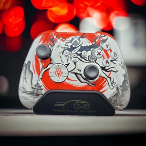 Może przedstawiać: Spersonalizowany kontroler Xbox z japońskim wzorem, przedstawiający białego wilka, czerwone akcenty i czarny stojak z napisem "BEETLE DASH". Kontroler ma szare drążki i kolorowe przyciski.