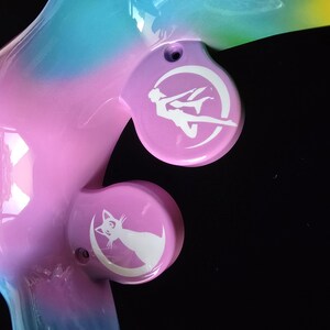 May include: A colorful, stylized sculpture with a gradient of pink, blue, and yellow hues. The sculpture features two circular, light purple accents with white illustrations of a figure and a cat on a crescent moon.