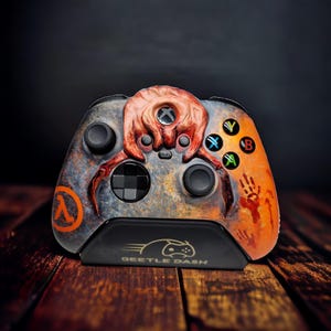 May include: Custom Xbox controller with a unique design. The controller has a rust-coloured and grey finish, with a detailed creature design on the top. The right side has an orange gradient with a handprint. The controller rests on a black stand with the text "Beetle Dash".