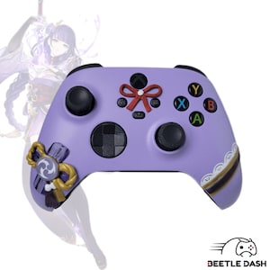 Puede incluir: Un mando de Xbox morado con botones negros y una cruceta. El mando presenta detalles decorativos, incluyendo un lazo rojo y adornos dorados. Las letras X, Y, A y B están en el lado derecho.