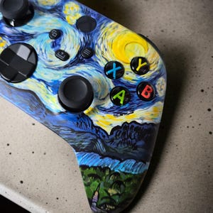 May include: Custom-painted video game controller with a starry night design in blue, yellow, and white. The controller has black buttons and joysticks, with coloured action buttons.
