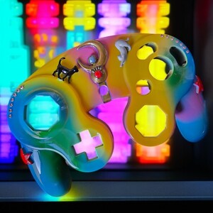 May include: A custom-painted video game controller with a gradient of yellow, green, and blue. It features a black cat figurine, a crescent moon, and jewel accents. The controller has a glowing blue light and a pink cross button.