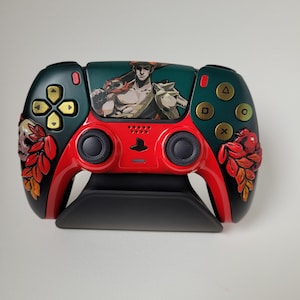 May include: Custom video game controller with a red and black base, featuring a character illustration and floral details. Gold buttons and a black stand complement the design. The controller has a dark green and red colour scheme.