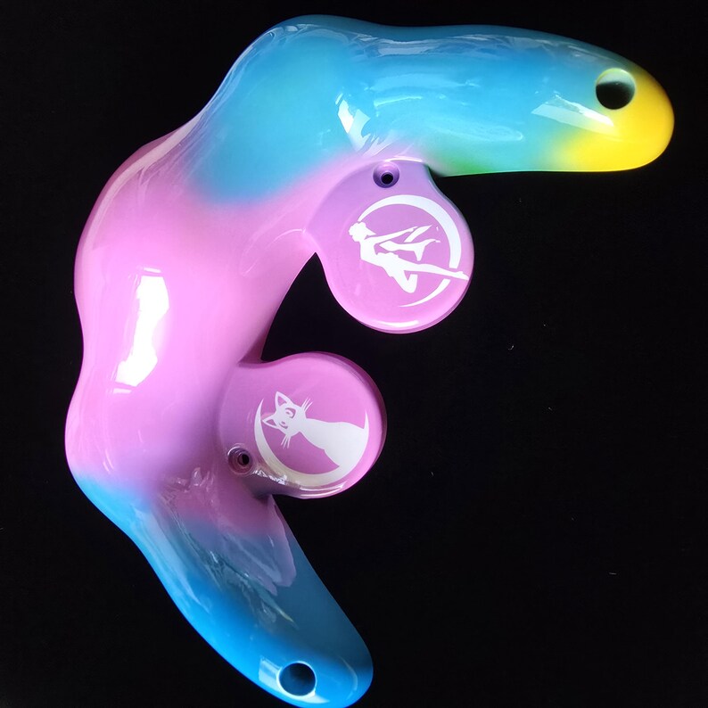 May include: A colorful, abstract-shaped object with a gradient of blue, pink, and yellow. It features two circular pink accents with white illustrations of a figure and a cat on a crescent moon. The object has a glossy finish and a hole at each end.