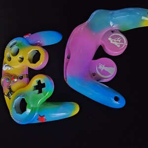 May include: Two custom-painted video game controller shells. One is yellow, green, and blue with a red star and a cross-shaped button. The other is pink, purple, and blue with white graphics. Both have a glossy finish.