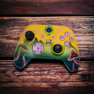 May include: Custom-painted video game controller with a yellow, green, and pink design. The controller features a landscape with a compass rose, a monster, and other graphic elements. The buttons are pink and yellow.