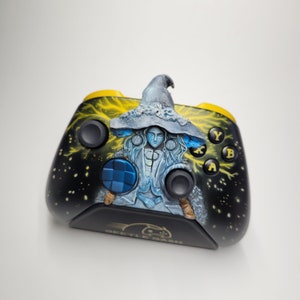 May include: Custom-painted black and yellow video game controller featuring a detailed illustration of a figure with a hat. The controller has a black base with yellow accents and buttons labeled Y, B, and A. The design includes a lightning bolt pattern.