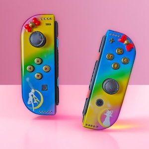 May include: Two custom Nintendo Switch Joy-Cons with a gradient rainbow design. Each controller features a red bow, black joystick, and gold buttons. One controller has a Sailor Moon graphic, the other a Luna cat graphic.