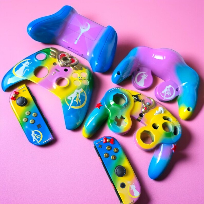 May include: A collection of colorful video game controllers in various designs. The controllers feature a gradient of colors, including blue, yellow, and purple, with character-themed artwork. The controllers are arranged on a pink surface.
