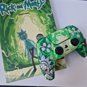 May include: A green and white camouflage-patterned video game controller with a Rick and Morty graphic. The controller is next to a book with the Rick and Morty logo and characters. The book and controller have a green color scheme.