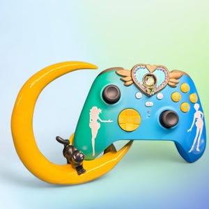 Può includere: Un controller per videogiochi personalizzato nei toni del blu e del verde acqua, ornato da un emblema a forma di cuore e dettagli alati. Il controller presenta una luna crescente gialla e una statuina di gatto nero. Il design include sagome.
