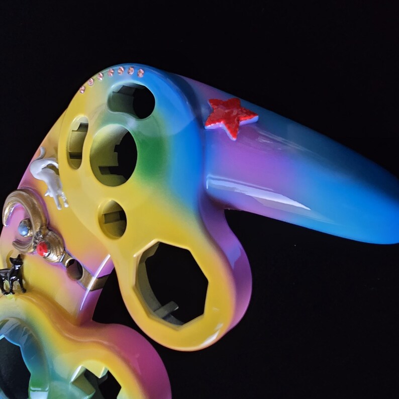 May include: A colorful, custom-painted game controller with a rainbow gradient design. The controller features a red star, small decorative charms, and a glossy finish. The design incorporates yellow, blue, purple, and pink hues.