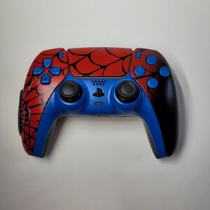 May include: A custom-designed video game controller with a Spider-Man theme. The controller features a red and black spiderweb design on the top, with blue accents on the buttons and handles. The controller has black joysticks and a PlayStation symbol.