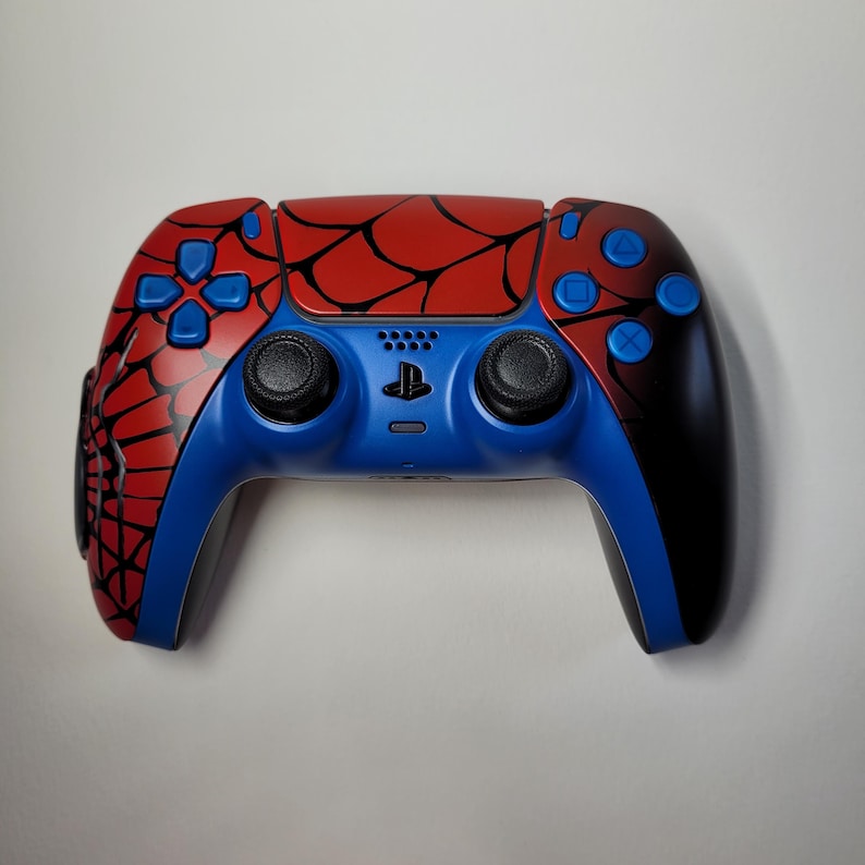May include: A custom-designed video game controller with a Spider-Man theme. The controller features a red and black spiderweb design on the top, with blue accents on the buttons and handles. The controller has black joysticks and a PlayStation symbol.