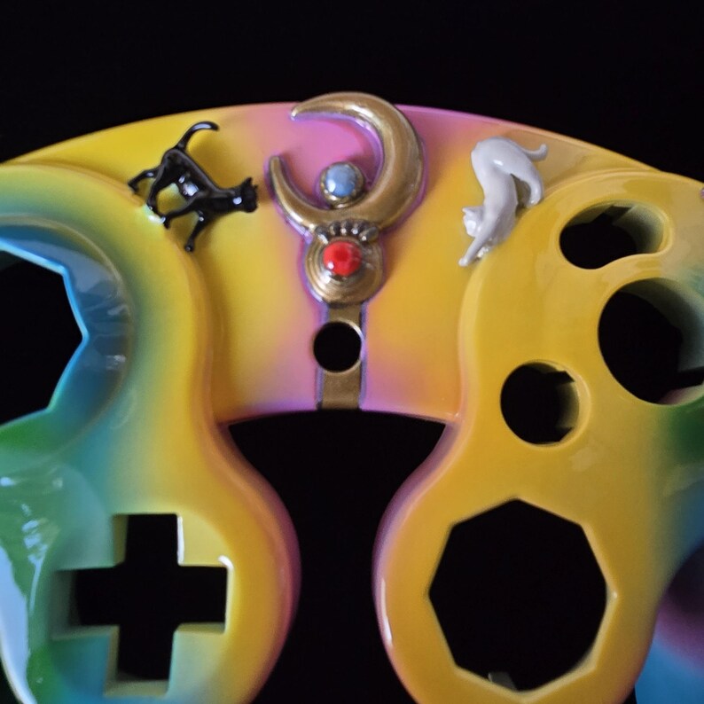 May include: A custom-painted video game controller with a rainbow gradient design. The controller features a gold crescent moon with a red jewel and two cat figurines. The buttons and directional pad are black.