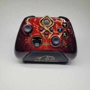 Könnte beinhalten: Individuell gestalteter Videospiel-Controller mit einem auffälligen rot-goldenen Farbschema. Der Controller verfügt über ein einzigartiges Design mit goldenen Akzenten und Symbolen, und der Text "DEETLE DASH" ist auf der Ladestation zu sehen.