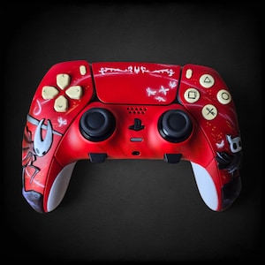 May include: A custom red games controller with white detailing and black joysticks. The controller features artwork of a character from the video game Hollow Knight. The buttons are white with symbols.