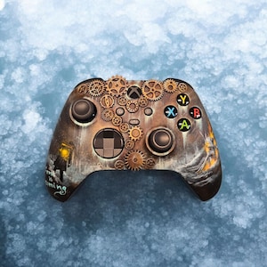 May include: A custom game controller featuring a weathered metallic finish with raised bronze-colored gear embellishments across the top. The surface displays a painted scene with a glowing lantern and the text Frost is coming in white script.