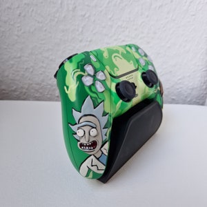 Puede incluir: Un mando de videojuegos verde y amarillo con un diseño de personaje de dibujos animados. El mando tiene joysticks y botones negros y está sobre un soporte negro. El diseño incluye un personaje de dibujos animados con pelo blanco y una bata de laboratorio.