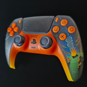 May include: Custom orange and gray PlayStation 5 controller. The controller features orange buttons and accents, with a gray top and a painted landscape design on the right side. The controller has a black background.
