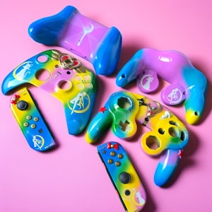 May include: A collection of colorful video game controllers in various designs. The controllers feature a gradient of colors, including blue, yellow, and purple, with character-themed artwork. The controllers are arranged on a pink surface.