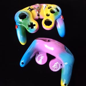 Puede incluir: Dos mandos de videojuegos pintados a medida. Los mandos están pintados con un degradado arcoíris de colores, incluyendo amarillo, verde, azul y rosa. Un mando tiene un símbolo de más y el otro una silueta de gato blanco.