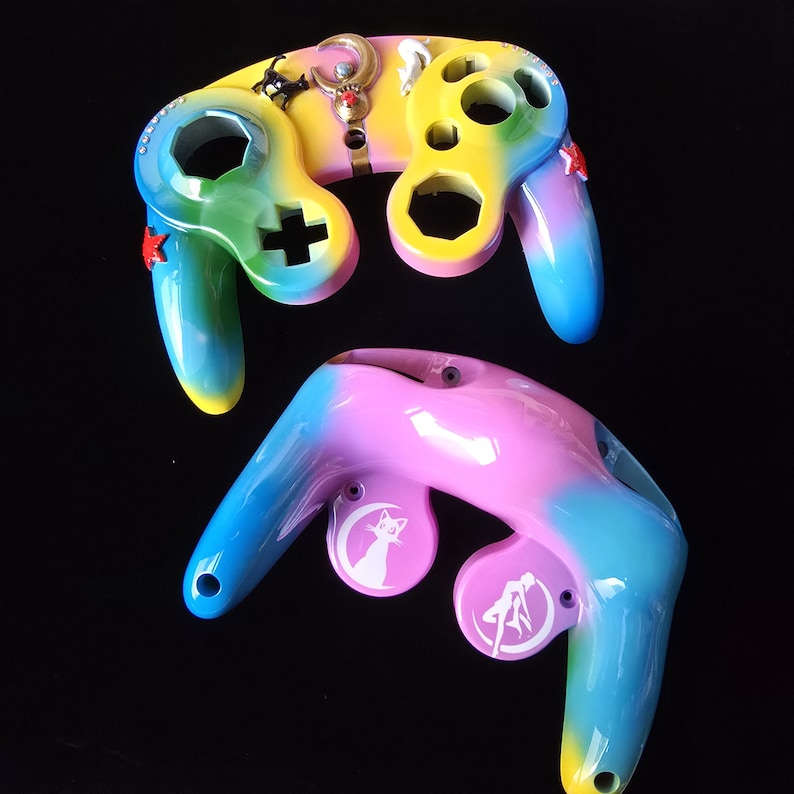 May include: Two custom-painted video game controllers. The controllers are painted with a rainbow gradient of colors, including yellow, green, blue, and pink. One controller has a plus symbol and the other has a white cat silhouette.