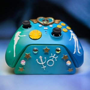 Puede incluir: Un mando de videojuegos personalizado en tonos azul y verde azulado, adornado con estrellas doradas, gemas y un emblema en forma de corazón. Cuenta con figuras de silueta blanca y botones amarillos. El mando tiene joysticks negros.
