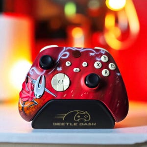 Può includere: Controller Xbox rosso e nero personalizzato con un design unico. Il controller presenta un'illustrazione stilizzata sul davanti, dettagli bianchi e una base nera con la scritta "BEETLE DASH". Ha joystick e pulsanti neri.