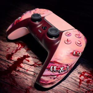 Puede incluir: Un mando de videojuegos personalizado con un diseño único. El mando presenta una combinación de colores rojo oscuro y rosa con un diseño gráfico que se asemeja a piel cosida. Los botones y joysticks son negros.
