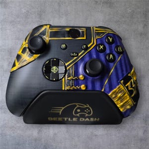 Op de afbeelding: Op maat gemaakte videogamecontroller met een zwart, blauw en goud kleurenschema. De controller heeft een uniek ontwerp met ingewikkelde details en de tekst "Beetle Dash" op de oplaadstandaard.
